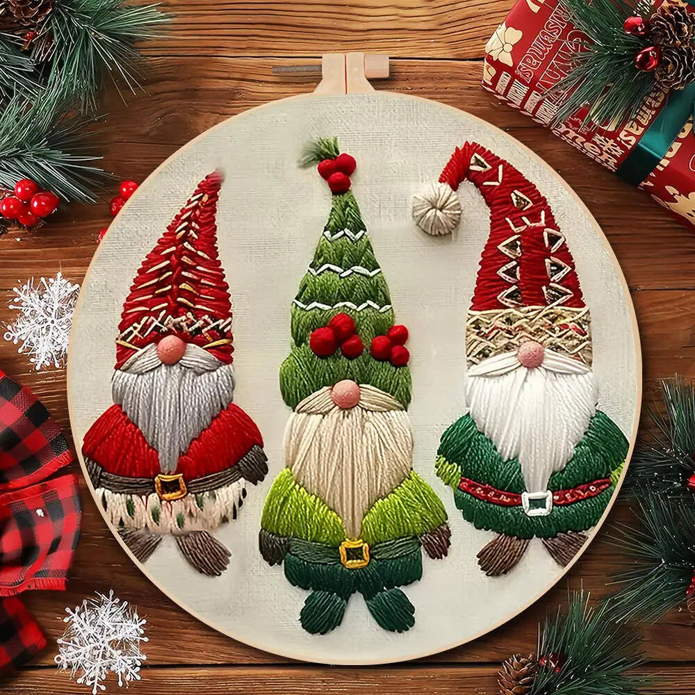 Embroidery hoop with three embroidered gnomes on a wooden surface with Christmas decorations.