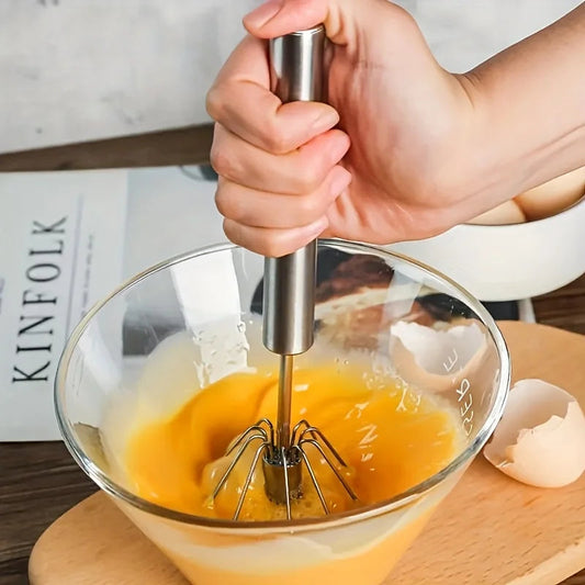 Hand using a manual egg beater to mix eggs in a glass bowl on a wooden surface.