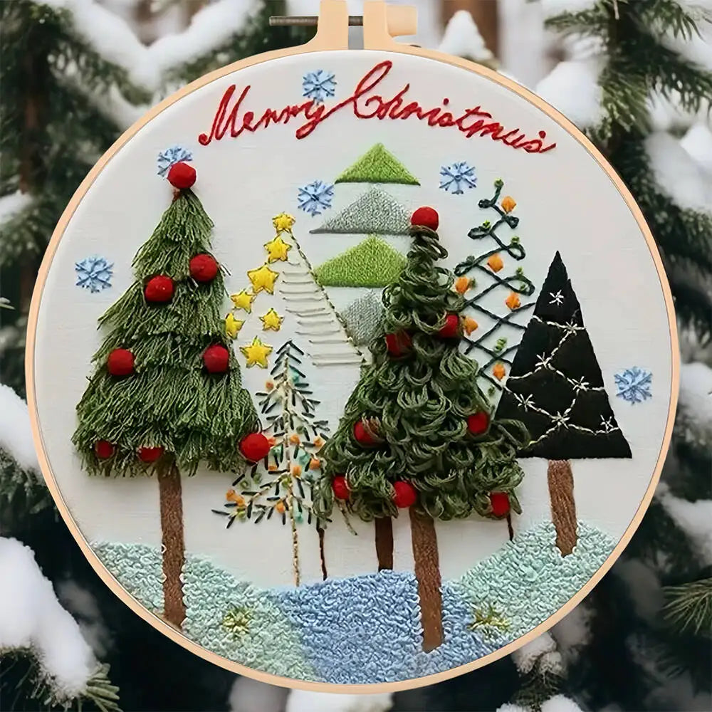 Embroidery hoop with Christmas-themed design featuring trees and 'Merry Christmas' text, surrounded by snowy trees.