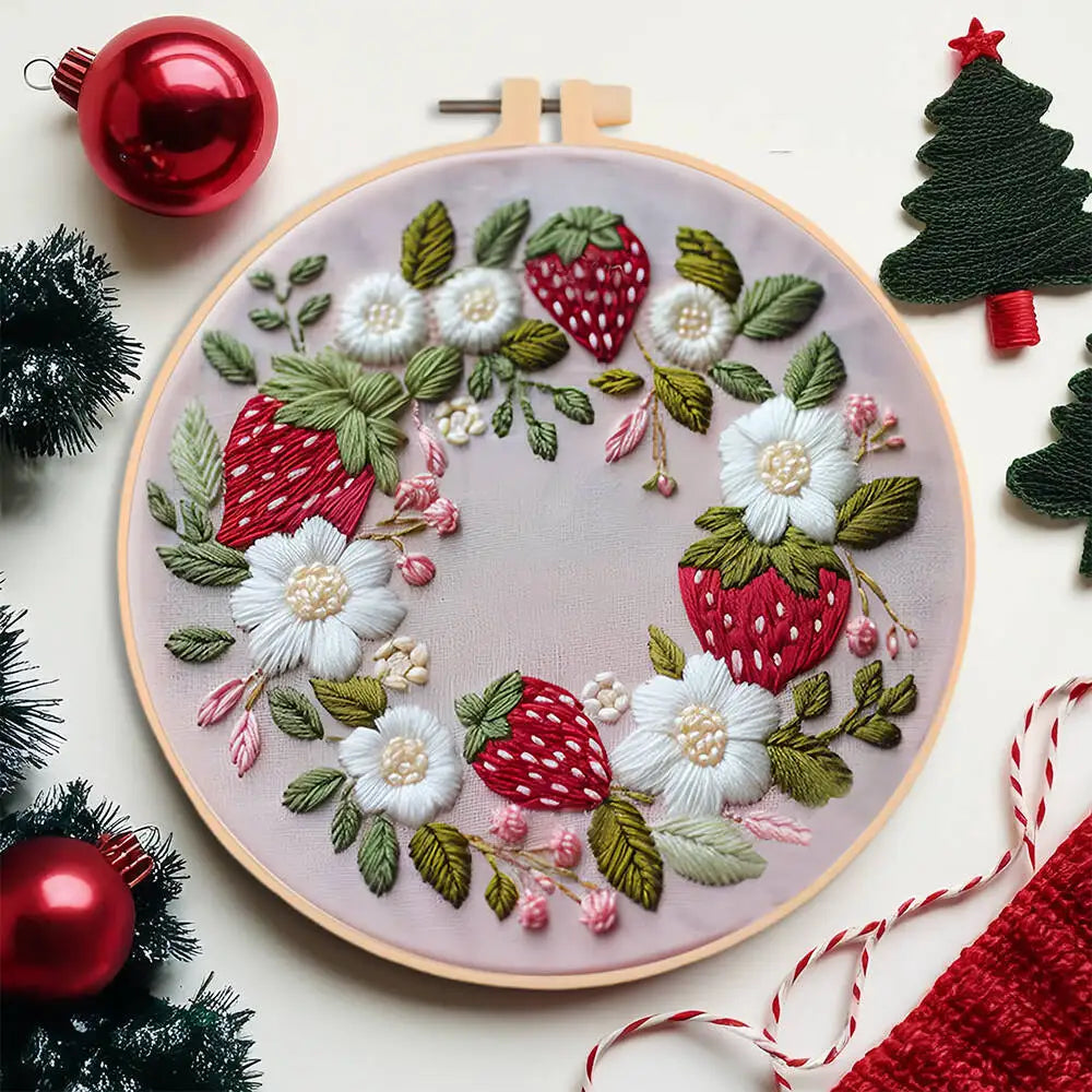 Embroidery hoop with strawberry and flower pattern surrounded by Christmas decorations on a white background