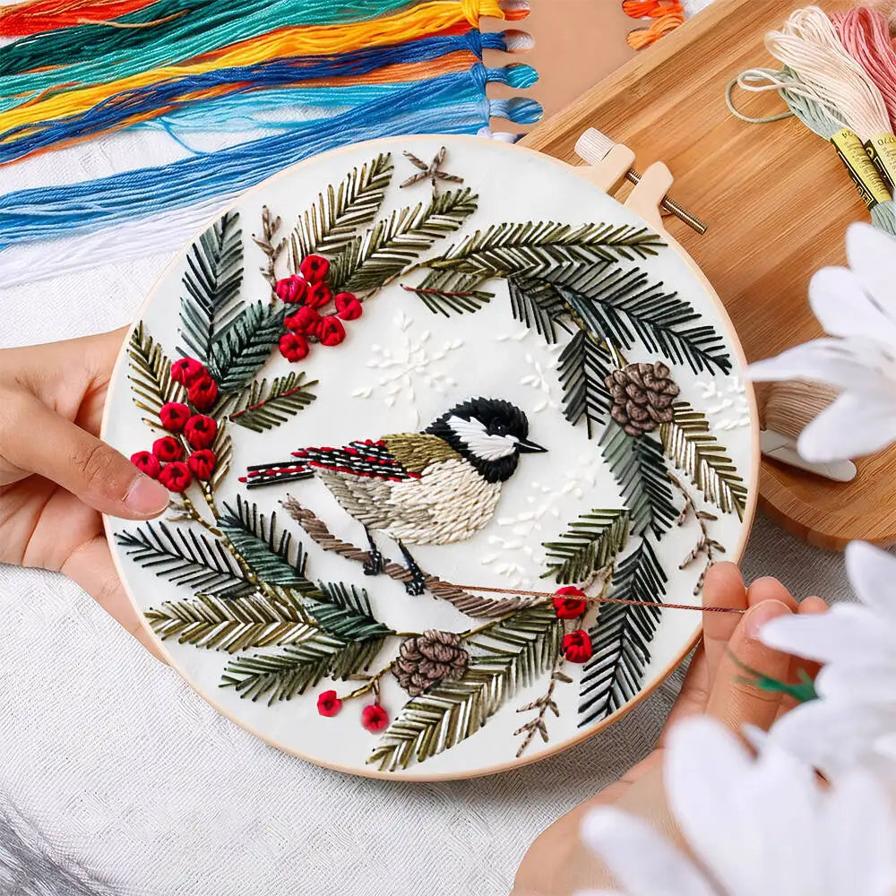 Embroidery hoop with a bird and winter-themed design held by hands, surrounded by embroidery threads.