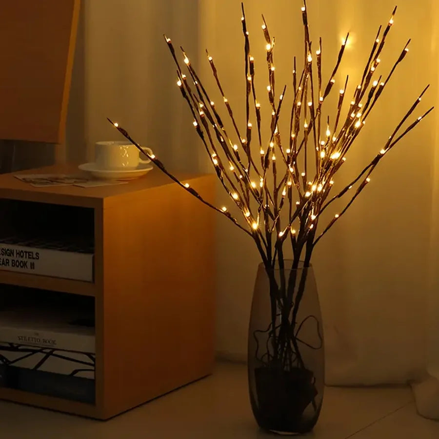 Decorative branches with lights in a vase on a table next to a bookshelf.