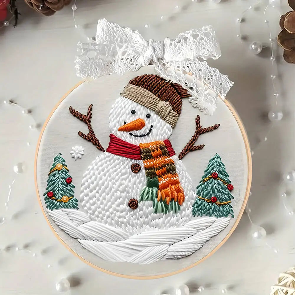 Embroidery hoop with a snowman design featuring a scarf, trees, and a bow on a textured white background.