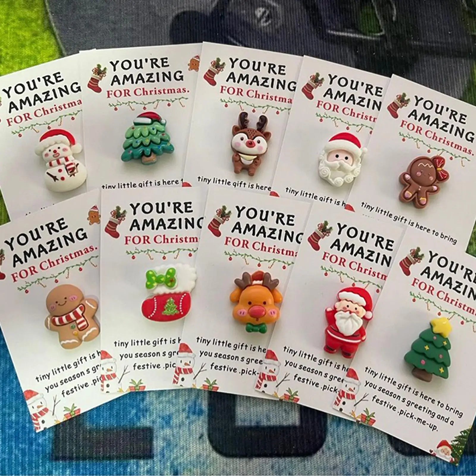 Multiple 'You're Amazing for Christmas' cards with small figurines on a blue surface.